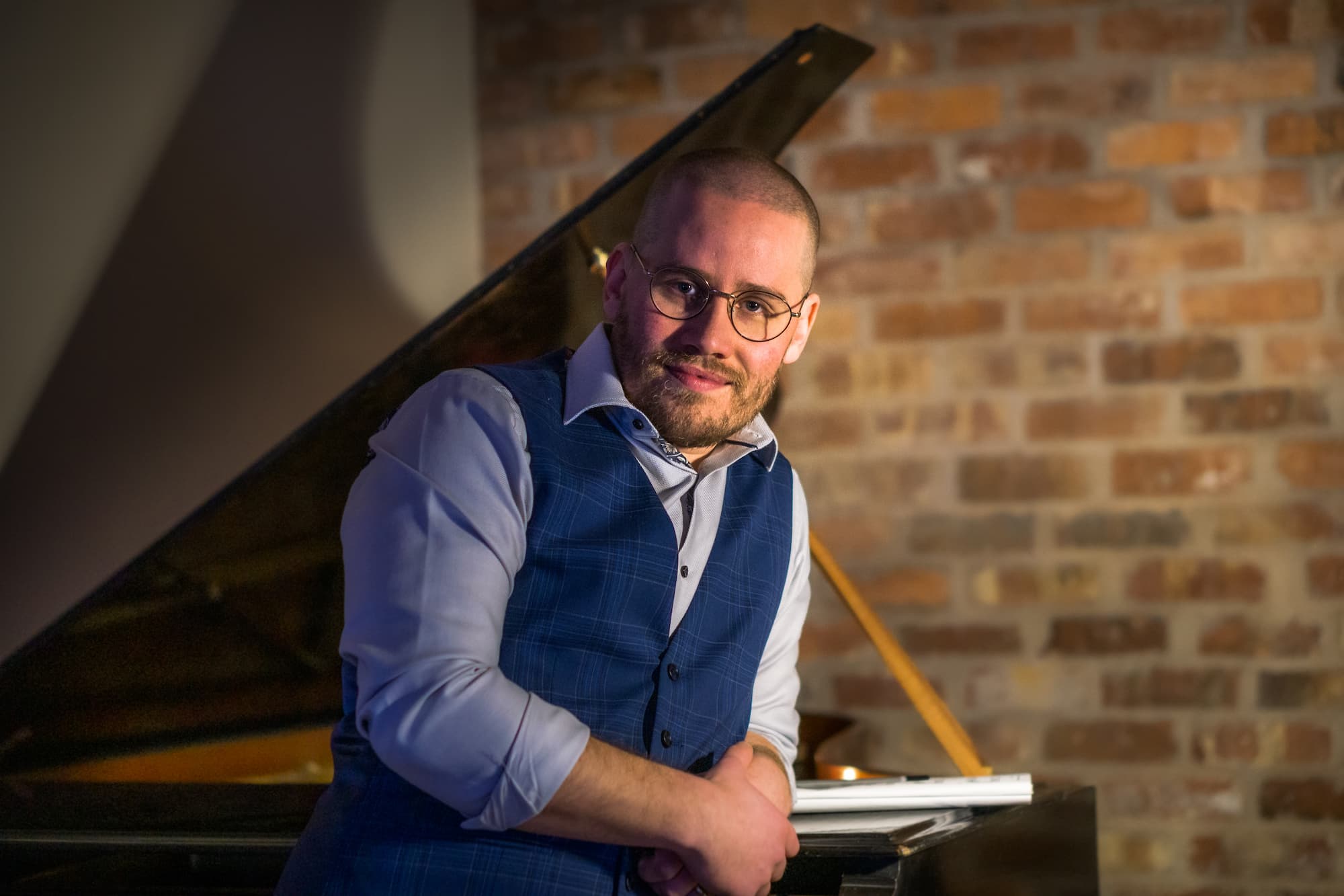 Man in glasses and blue vest leaning against a grand piano with brick wall background.